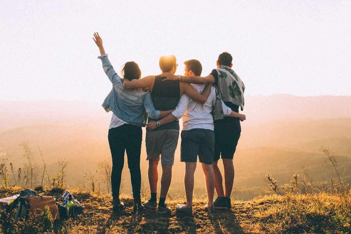 Four friends standing arm in arm on a hilltop at sunset, seen from behind