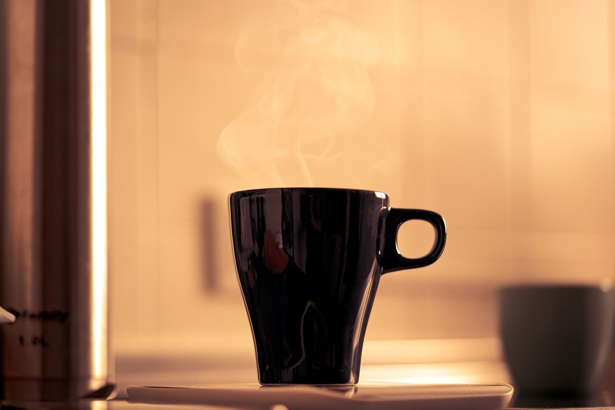 A black glazed mug steaming on a saucer, lit by a warm kitchen behind it