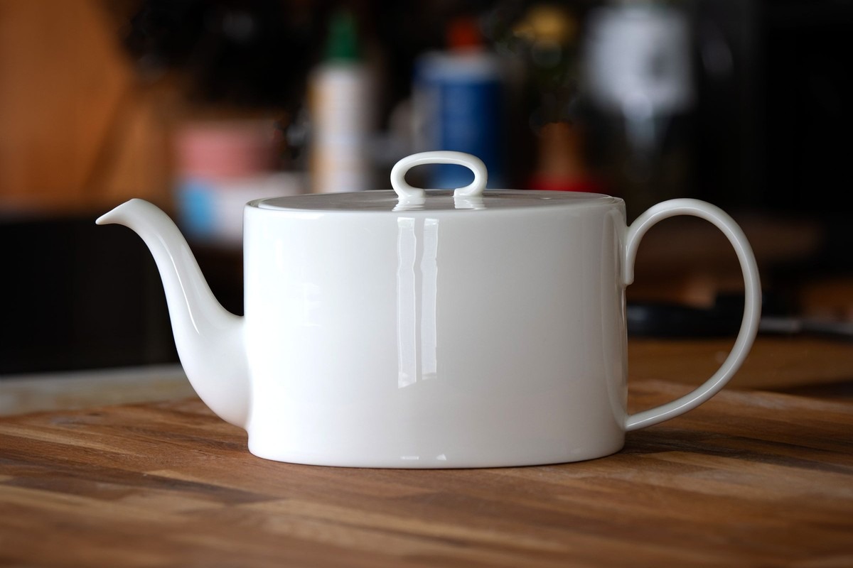 A white porcelain teapot on a wooden kitchen table, a cluttered worktop out of focus behind it