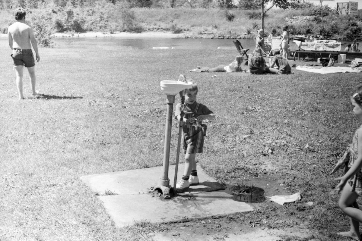 A black and white photo of a riverside park, a child at a drinking fountain