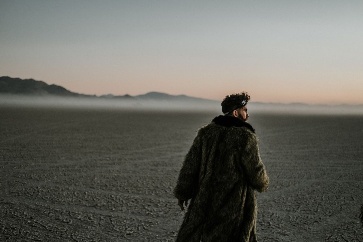 A man in a heavy fur coat and fur hat standing on a misty flat desert at dusk, seen from behind