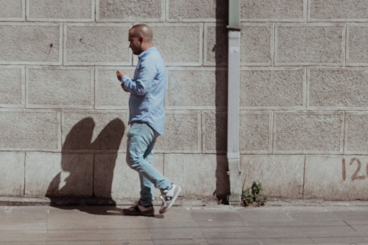 A man walking along a plain block wall, casting a hard shadow across it
